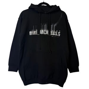 Puede incluir: Sudadera negra con bolsillo delantero y capucha. La sudadera presenta el texto "NINE INCH NAILS" en blanco y con un diseño estilizado en el pecho. El diseño incluye líneas verticales y un elemento gráfico curvo.