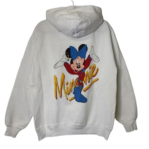 Puede incluir: Sudadera con capucha blanca con un gr&aacute;fico colorido de Minnie Mouse en la espalda. El gr&aacute;fico incluye a Minnie Mouse con un atuendo rojo y un lazo azul, y la palabra "Minnie" en letras amarillas.
