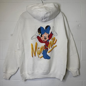 Puede incluir: Sudadera con capucha blanca con un gr&aacute;fico colorido de Minnie Mouse en la espalda. Minnie lleva un lazo azul, una chaqueta roja y pantalones azules. La palabra "Minnie" est&aacute; escrita en amarillo.