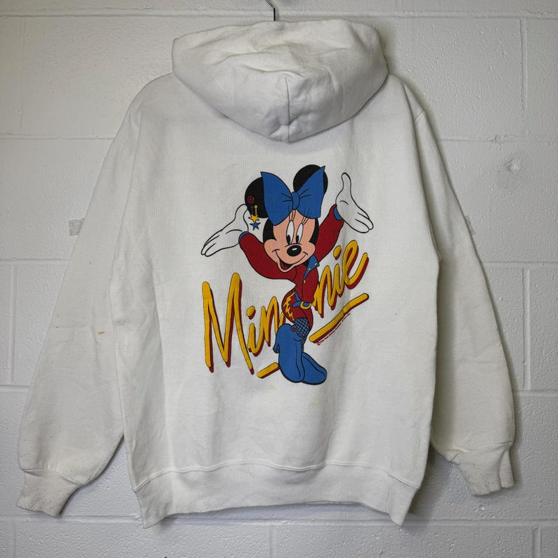 Puede incluir: Sudadera con capucha blanca con un gr&aacute;fico colorido de Minnie Mouse en la espalda. Minnie lleva un lazo azul, una chaqueta roja y pantalones azules. La palabra "Minnie" est&aacute; escrita en amarillo.