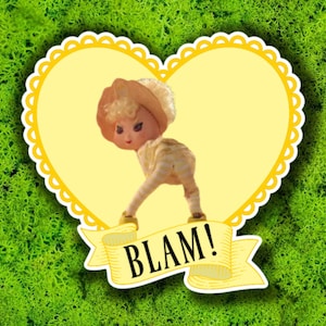 May include: A vintage doll with a hat and striped outfit is featured in a yellow heart-shaped frame with a scalloped edge. The doll is bent over with the word "BLAM!" on a banner below, set against a green moss background.