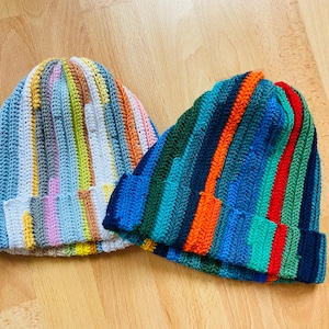 Könnte beinhalten: Zwei gestreifte Häkelmützen. Eine Mütze ist blau, grün, orange und rot. Die andere Mütze ist weiß, blau, gelb, rosa und braun.