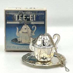 Könnte beinhalten: Ein silberfarbener Tee-Ei-Einsatz in Form einer Miniatur-Teekanne, der auf einer passenden kleinen Untertasse mit einer dünnen Kette ruht. Das Produkt wird vor seiner Originalverpackung aus Karton mit der Aufschrift TEE-EI präsentiert.