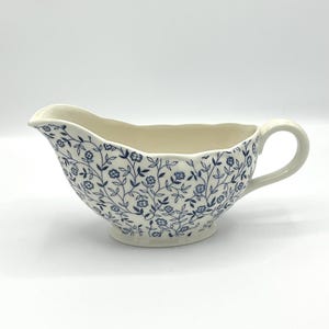 Könnte beinhalten: Cremefarbene Sauciere mit blauem Blumenmuster. Das Boot hat einen gewellten Rand, einen Griff und einen Ausguss zum einfachen Ausgießen. Das florale Design bedeckt die Außenseite des Bootes.