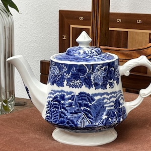 Könnte beinhalten: Eine weiße Keramik-Teekanne mit einem blauen Transferdruck-Muster, das eine ländliche Landschaft mit Häusern und Bäumen zeigt. Die Kanne hat einen passenden Deckel mit spitzem Knauf, einen geschwungenen Ausguss und einen verzierten Henkel.
