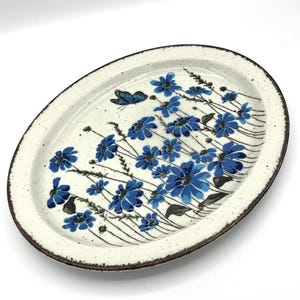 Könnte beinhalten: Ovale Servierplatte aus Keramik mit cremefarbenem Grund und braunem Rand. Sie ist mit blauen Blumen und einem blauen Schmetterling verziert. Die Blumen sind in einem fließenden Muster über die Oberfläche angeordnet.