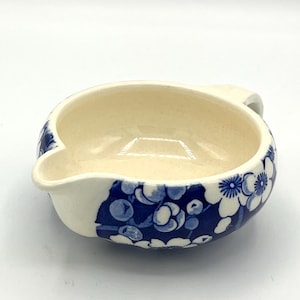 Könnte beinhalten: Cremefarbene Keramikschale mit blauem Blumenmuster. Die Schale hat einen Ausguss und einen Griff. Das blaue Blumendesign befindet sich auf der Außenseite. Das Innere ist cremefarben und glänzend.