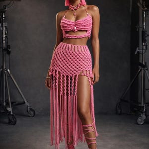 Op de afbeelding: Een roze gehaakte set met een haltertop, een rok met hoge taille en een hoge split, en bijpassende accessoires. De rok heeft lange franjes. De outfit wordt gecombineerd met sandaaltjes met bandjes en een hoofdband.