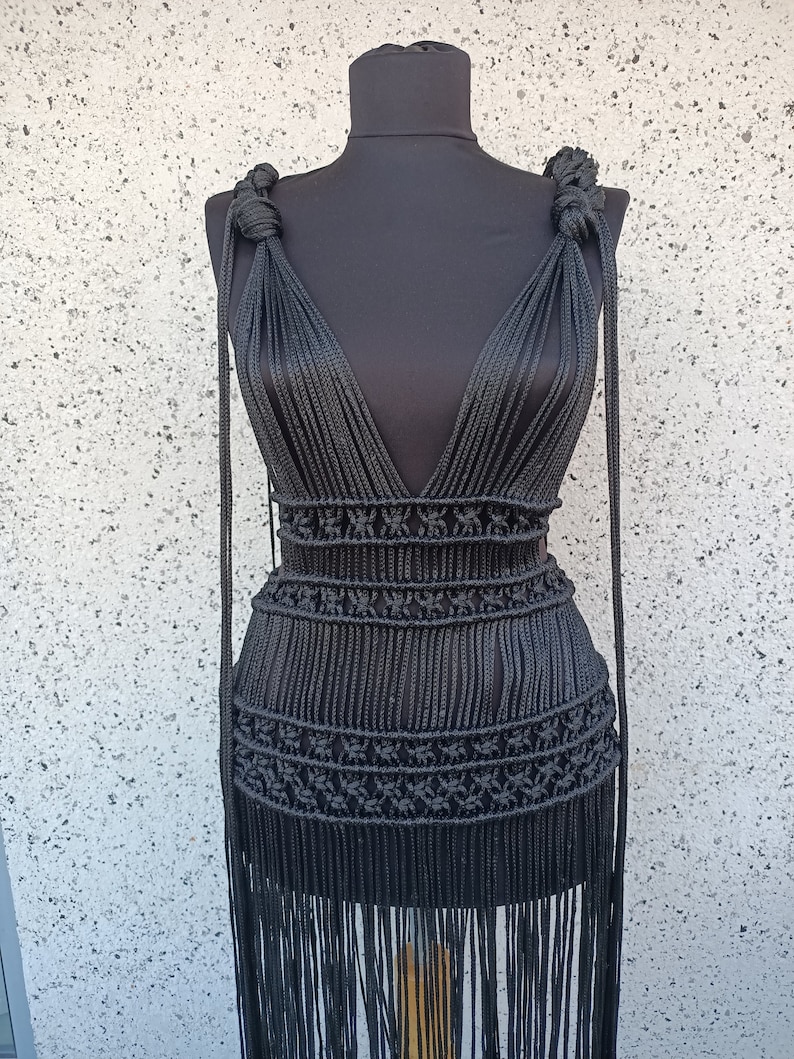 May include: A black crocheted dress with a deep V-neckline and fringe detailing. The dress is made with thick black cord and has a fitted bodice and a flowing skirt.