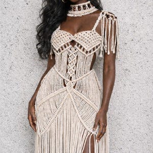 以下が含まれることがあります： ベージュのマクラメドレス。ハイウエストのスリット、フリンジのディテール、お揃いのチョーカーが特徴です。ドレスは織りのボディスとストラップ、流れるようなスカートが特徴です。夏のイベントにぴったりのボヘミアンスタイルです。