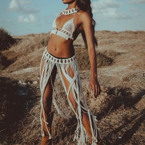 May include: A woman wearing a white crocheted top and skirt with fringe. The skirt is tied at the waist with a brown beaded belt. The woman is standing in a field of long grass.
