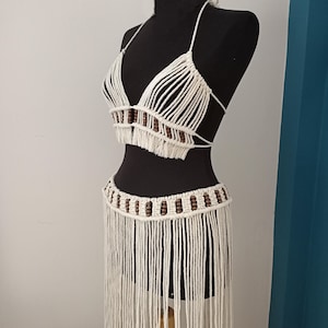 Puede incluir: Un conjunto de top y falda de crochet blanco con cuentas de madera marrón. El top tiene forma triangular y la falda tiene flecos.