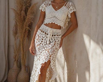 May include: A cream-colored crochet two-piece set. The top has a V-neck and short sleeves. The skirt is floor-length with a high slit. The set is made of a delicate, open-weave crochet pattern, perfect for a bohemian or beach-inspired look.