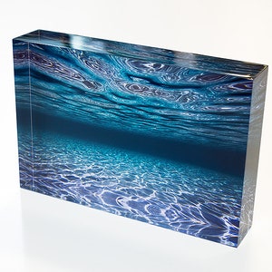 May include: A close-up of a blue and white abstract print of rippling water. The print is on a clear acrylic block.