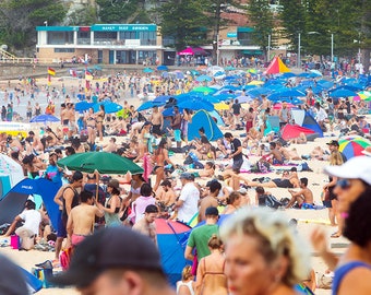 May include: A crowded beach with people sitting and standing under colorful umbrellas and canopies. There are many people in the water and on the sand. The beach is surrounded by buildings and trees.