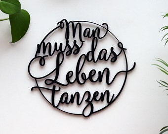 May include: Black wooden wall art with the German phrase "Man muss das Leben tanzen" (One must dance life) in a cursive font, enclosed in a circular frame. The art is set against a white background with green plants.