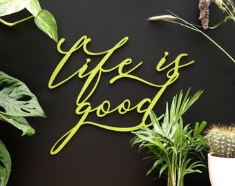 May include: A yellow wooden sign with the words "Life is good" written in cursive script. The sign is hanging on a black wall with green plants around it.