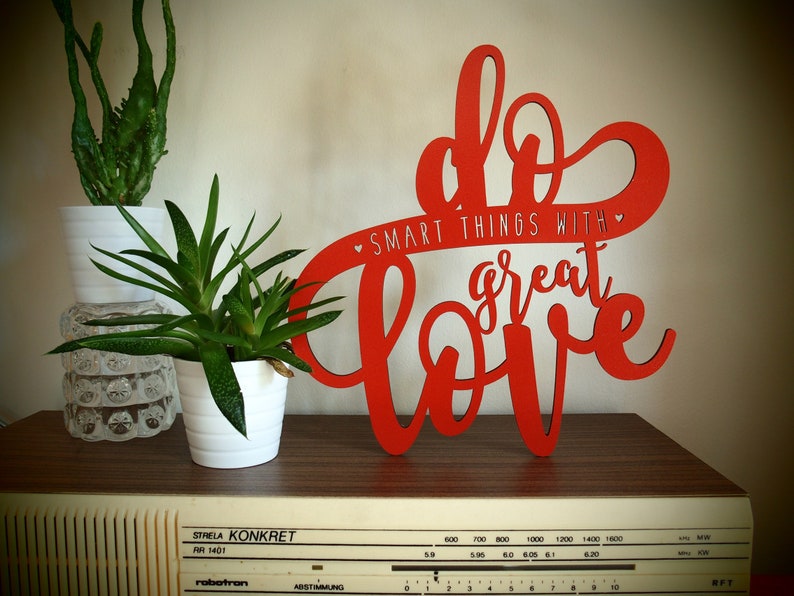 May include: Red wooden sign with the text "Do smart things with great love" in a decorative font. The sign is on a wooden surface with a radio below it.