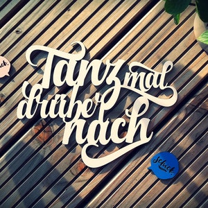 Könnte beinhalten: Ein weißes Holzschild mit dem Text "Tanz mal drüber nach" in einer dekorativen Schreibschrift. Das Schild liegt auf einer Holzoberfläche mit zwei kleinen Sprechblasen, die "Schick" in rosa und blau sagen.