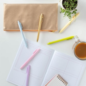 May include: A flat lay of a white surface with a gold pencil case, four pens, a small notepad, a larger notebook, a cup of coffee, a small potted plant, and a blue box with push pins.