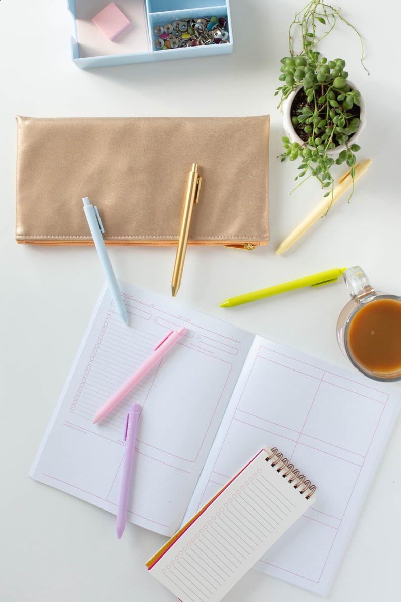May include: A flat lay of a white surface with a gold pencil case, four pens, a small notepad, a larger notebook, a cup of coffee, a small potted plant, and a blue box with push pins.