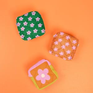 May include: Three square quilted pouches with zipper closures. One pouch is green with a pink flower pattern, one is brown with a pink flower pattern, and one features a color-blocked design in pink, orange, and lime green with a single large pink flower.