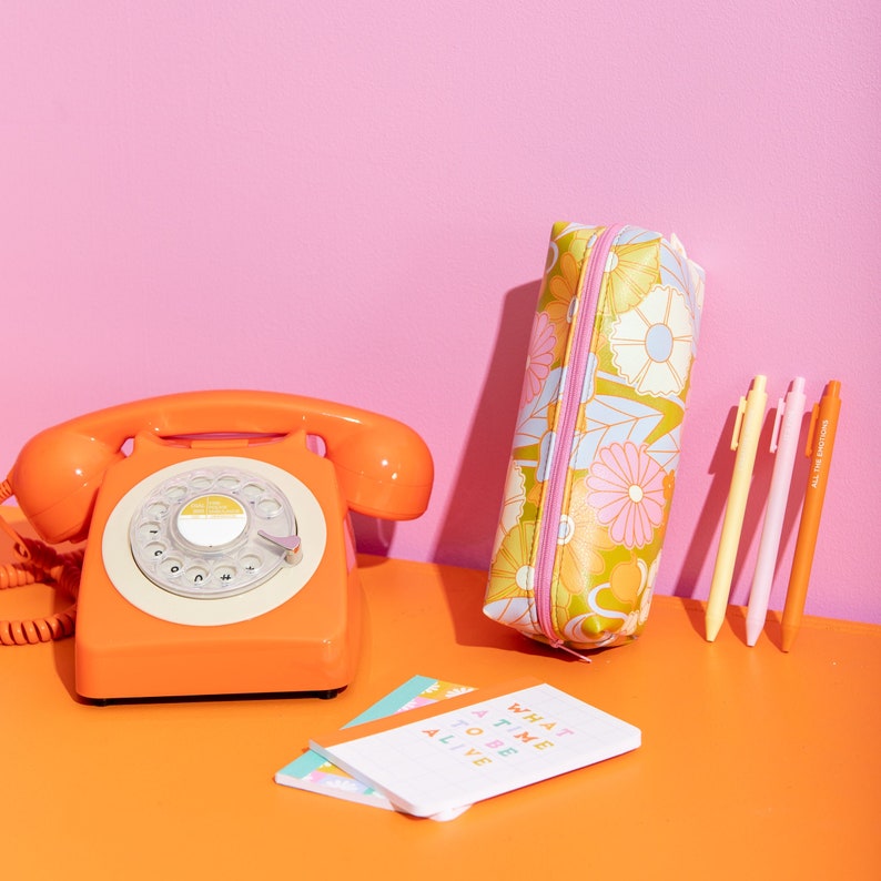 Gathering flowers jitterbug along with jotter pens and misc items. Pink background and an orange surface.