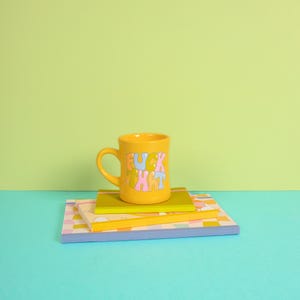 May include: Yellow ceramic mug with the phrase "F*CK THAT" in colorful letters, sitting on top of a stack of colorful notebooks.