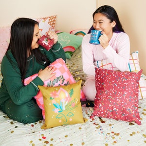 May include: Two people in pajamas are seated on a bed surrounded by decorative pillows and mugs. The pillows feature various designs, including a floral face, a checkered pattern, and the words "Merry" and "Fa La F*cking La". The mugs have holiday-themed text.