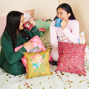 May include: Two people are sitting on a bed with pillows and a blanket. They are holding mugs and smiling. The pillows have festive designs with the text "Merry Christmas" and "La la la, it's Christmas".