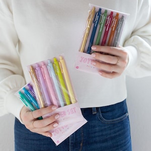 May include: Two sets of colorful pens with motivational phrases. The first set is a pack of six pastel pens with the phrase "The Power Set" and the second set is a pack of six metallic pens with the phrase "Jot It Down" and the text "Pack of Six".