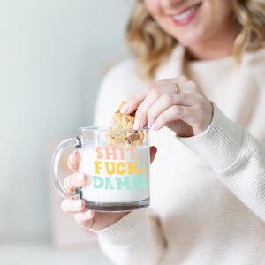 May include: A clear glass mug with a handle, filled with milk, and a cookie being dipped into it. The mug has the words "SHIT. FUCK. DAMN." in colorful letters. The cookie is a golden brown color.
