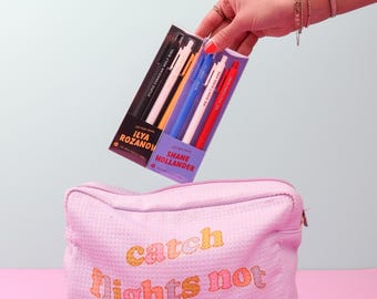 May include: A light pink zippered pouch with the text "catch flights not feelings" in orange and pink letters. Above the pouch, two sets of multi-colored jotter pens are held in their respective black and purple cardboard packaging boxes.