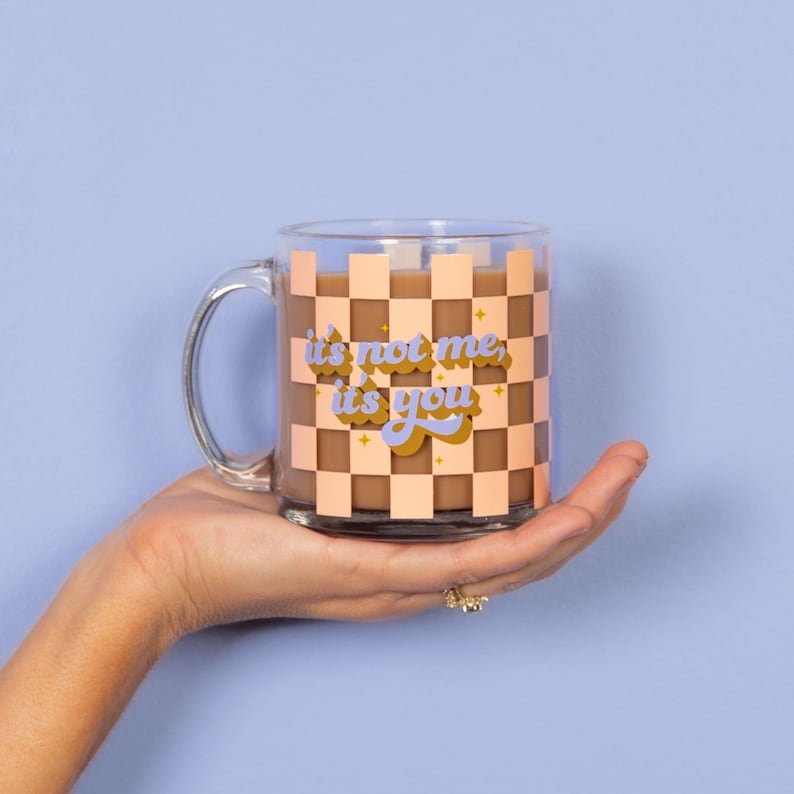 Puede incluir: Una taza de vidrio transparente con un patr&oacute;n a cuadros en marr&oacute;n y rosa. La taza tiene un asa y dice "it's not me, it's you" en una fuente retro con acentos de purpurina dorada.