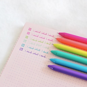 May include: A pink notebook with a dotted pattern and five colorful pens. The pens are pink, orange, yellow, green, and purple. The text "I said what I said" is written on the notebook in black ink.