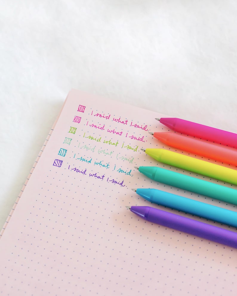 May include: A pink notebook with a dotted pattern and five colorful pens. The pens are pink, orange, yellow, green, and purple. The text "I said what I said" is written on the notebook in black ink.