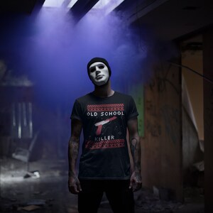 May include: A person wearing a black t-shirt with a red and white design that says "Old School Killer" and a white mask. The person is standing in a dark, abandoned building with blue smoke in the background.