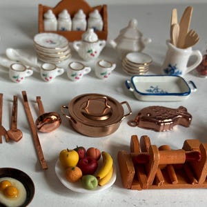 May include: A collection of miniature kitchen items, including a copper pot, frying pan with a fried egg, and a plate of fruit. Other items include a teapot, cups, plates, and spice jars. The items are arranged on a white surface.