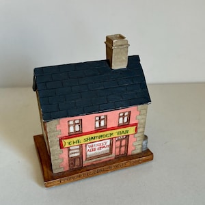 Può includere: Un modello in miniatura di un pub irlandese tradizionale chiamato "The Shamrock Bar". L'edificio è rosa con un tetto nero e una base marrone. Il cartello sopra la porta recita "The Shamrock Bar" e la finestra mostra "Whiskey Ales Stout".