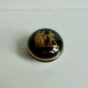 May include: A black and gold round trinket box with a gold image of three figures on the lid. The box is inscribed with the text "Art A".
