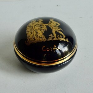 May include: A small, round, black ceramic trinket box with a gold trim and a gold image of three figures on the lid. The box is inscribed with the word "Corfu" in gold lettering.
