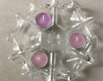 May include: Clear glass candle holder with a snowflake design, holding three pink candles. The candle holder has a textured surface and a starburst shape. The candles are round and have a white wick.