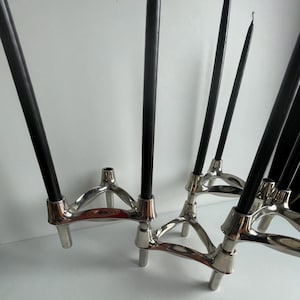 Può includere: Un set di candelabri in metallo argentato con candele nere. Ogni portacandela ha una base a treppiede e una parte superiore circolare per tenere la candela. Le candele sono alte e sottili, creando un'estetica moderna.