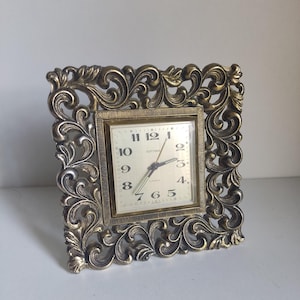 May include: A vintage square clock with a gold-colored, ornate frame. The clock face is cream-colored with black numerals and hands. The clock reads "ESTYMA" and has a classic, decorative design.