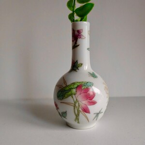 May include: A small white porcelain vase with a floral design featuring pink and green flowers. The vase has a round body and a narrow neck. A green leafy stem is in the vase.