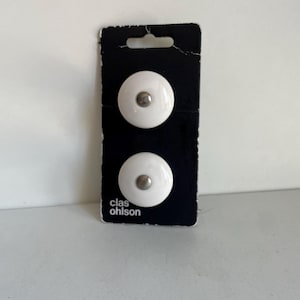 Pode incluir: Dois puxadores de armário de cerâmica branca com centros prateados são exibidos em um cartão preto. O cartão tem um furo na parte superior para pendurar e o texto "clas ohlson" impresso em branco.