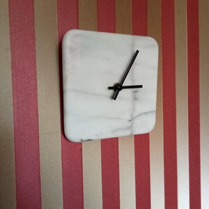 May include: Square white marble clock with black hands. The clock is mounted on a wall with vertical stripes in red and gold.