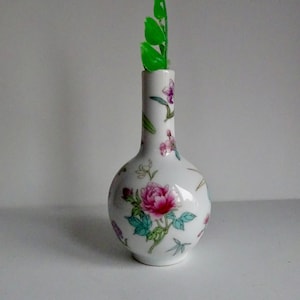 May include: A small white porcelain vase with a floral pattern in shades of pink, green, and blue. The vase has a round body and a narrow neck. A green leaf is placed in the vase.