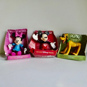 Könnte beinhalten: Drei Disney-Charakter-Figuren in ihrer Verpackung. Mickey Mouse trägt eine rote Hose und gelbe Schuhe. Minnie Mouse trägt ein rosa Kleid und gelbe Schuhe. Pluto ist ein gelber Hund mit einem grünen Halsband. Jede Schachtel trägt den Namen des Charakters und "Een echte Disney Original" aufgedruckt.