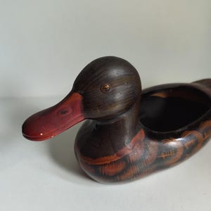 Könnte beinhalten: Holz-Entenfigur mit dunkelbraunem Körper und Kopf, rotbraunen Akzenten auf den Flügeln und einem roten Schnabel. Die Ente hat ein kleines, offenes Fach auf dem Rücken, was darauf hindeutet, dass sie ein dekorativer Behälter sein könnte.